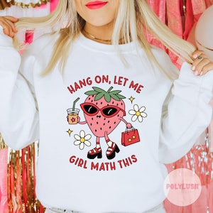 以下が含まれることがあります： 白いスウェットシャツに、サングラスをかけてショッピングバッグを持った漫画のイチゴが描かれています。スウェットシャツには「Hang on, let me girl math this」という文字がプリントされています。