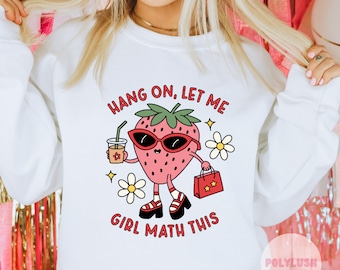 May include: White sweatshirt with a cartoon strawberry wearing sunglasses and holding a shopping bag. The text "Hang on, let me girl math this" is printed on the sweatshirt.