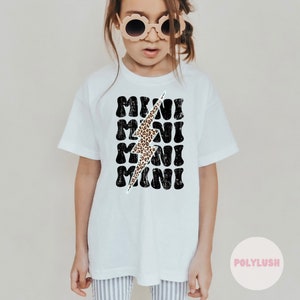 Könnte beinhalten: Weißes T-Shirt mit einer schwarz-weißen Grafik des Wortes "MINI", das viermal wiederholt wird, mit einem Blitz in Leopardenmuster in der Mitte.