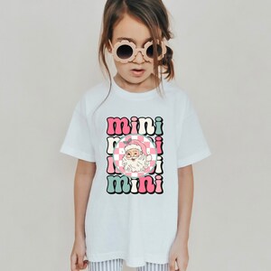 Könnte beinhalten: Weißes T-Shirt mit einem pinkfarbenen, grünen und weißen Grafikdesign mit dem dreimal wiederholten Wort "mini" und einem Cartoon-Weihnachtsmann, der einen pinkfarbenen und weißen karierten Hut trägt.
