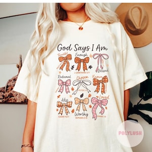 May include: Cream-colored t-shirt with the text "God Says I Am" and various words like "Thankful" and "Enough" arranged around colorful bow illustrations. The shirt also features Bible verses.