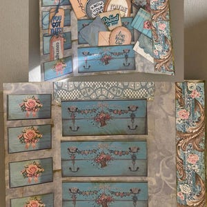 May include: A multi-layered paper craft project featuring blue drawer-themed pockets decorated with pink roses. The pockets contain various tags and circular cutouts with text including "You are smart," "Know your worth," "You're," "I'm proud of you," "Live laugh love," "You got this," and "Courage."