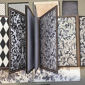 May include: A multi-page scrapbook or photo album featuring various black, white, and grey patterned pages. The designs include black and white diamonds, intricate damask swirls, vertical stripes, and floral motifs, all bound with dark brown edges.