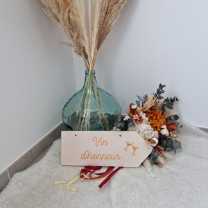 Peut inclure: Une pancarte en bois avec le texte "Vin d'honneur" en lettres orange et un graphique de deux verres de champagne. La pancarte est posée sur une surface blanche et moelleuse avec un bouquet de fleurs séchées et un vase de pampas dans le fond.