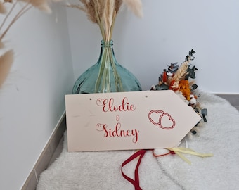 Könnte beinhalten: Ein hölzernes Pfeilschild mit dem Text "Elodie & Sidney" in roter Kursivschrift und zwei roten Herzen. Das Schild steht auf einem weißen, flauschigen Teppich.