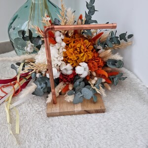 Op de afbeelding: Gedroogd bloemenboeket in tinten oranje, wit en rood, gerangschikt in een koperen standaard met een houten basis.