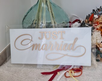 Può includere: Un cartello in acrilico bianco con le parole "JUST married" in lettere dorate. Il cartello è appoggiato su una superficie bianca e soffice con nastri rossi, gialli e bianchi.