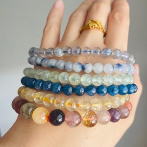 May include: A hand displays six beaded bracelets in various colors. The bracelets feature translucent and opaque beads in shades of blue, green, yellow, and purple. A gold ring is visible on the hand.