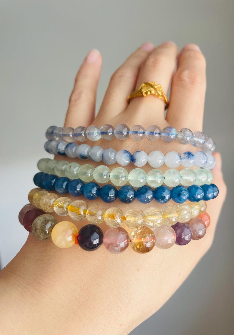 May include: A hand displays six beaded bracelets in various colors. The bracelets feature translucent and opaque beads in shades of blue, green, yellow, and purple. A gold ring is visible on the hand.