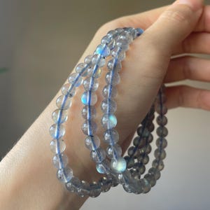 May include: A set of three beaded bracelets made of translucent, round, gray beads with blue flashes. The beads are strung on a blue elastic cord. The bracelets are held in a hand, showcasing the jewelry's design and color.