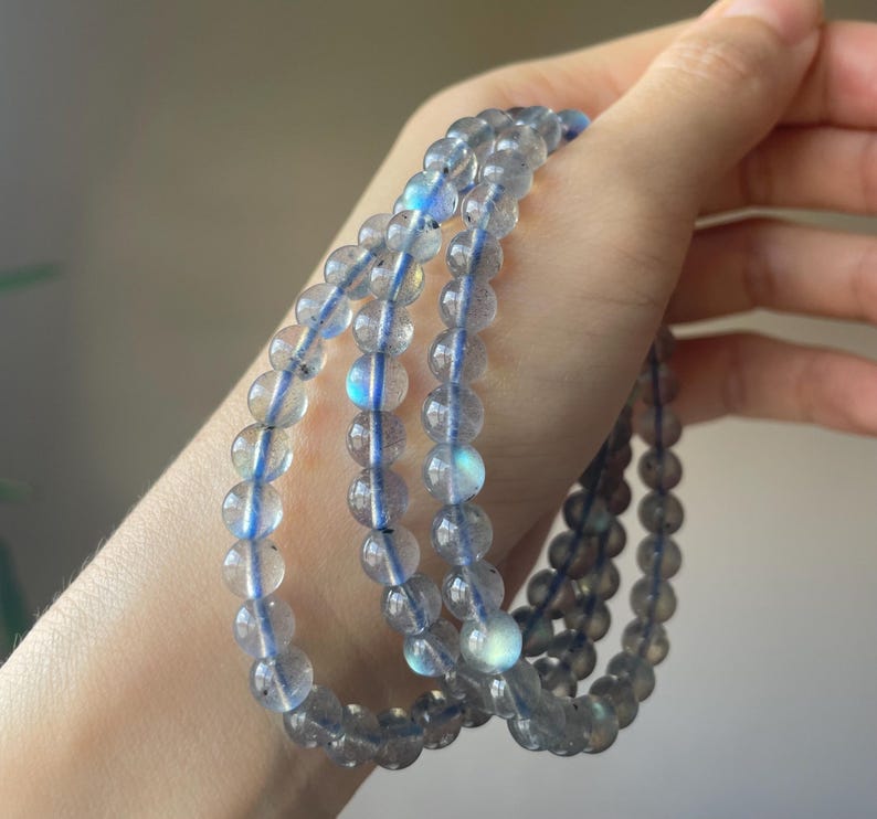 May include: A set of three beaded bracelets made of translucent, round, gray beads with blue flashes. The beads are strung on a blue elastic cord. The bracelets are held in a hand, showcasing the jewelry's design and color.