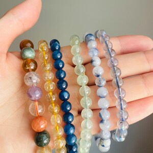 May include: A hand displays seven beaded bracelets in various colors. The bracelets feature round beads in shades of brown, yellow, blue, green, and clear. Each bracelet is strung on a clear or yellow elastic cord, showcasing the natural colors of the gemstones.