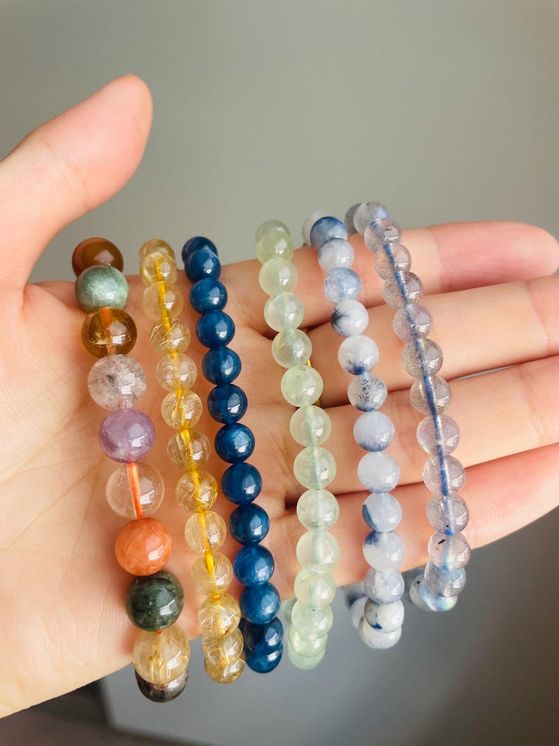 May include: A hand displays seven beaded bracelets in various colors. The bracelets feature round beads in shades of brown, yellow, blue, green, and clear. Each bracelet is strung on a clear or yellow elastic cord, showcasing the natural colors of the gemstones.
