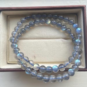 May include: Two strands of beaded bracelets with translucent, round, gray-blue beads. The beads are strung on a blue elastic cord. The bracelets are displayed inside a brown jewelry box with a cream-colored interior.
