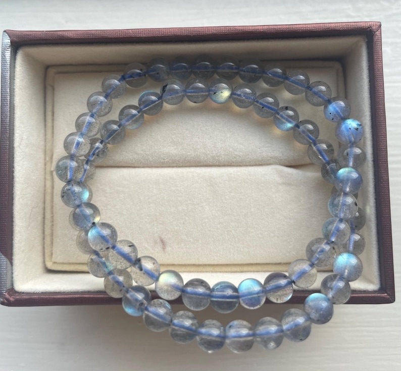 May include: Two strands of beaded bracelets with translucent, round, gray-blue beads. The beads are strung on a blue elastic cord. The bracelets are displayed inside a brown jewelry box with a cream-colored interior.