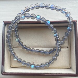 May include: Two beaded bracelets made of round, translucent gray beads with flashes of blue and iridescent color. The bracelets are displayed in a brown jewelry box with a beige interior.