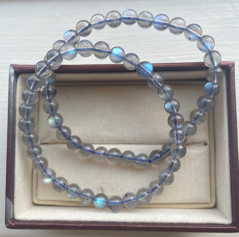 May include: Two beaded bracelets made of round, translucent gray beads with flashes of blue and iridescent color. The bracelets are displayed in a brown jewelry box with a beige interior.