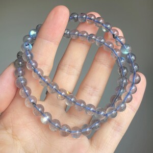 May include: Two beaded bracelets with clear, round beads and a blue string. The beads have a subtle iridescent quality, reflecting shades of blue and gray. The bracelets are displayed against a neutral background.