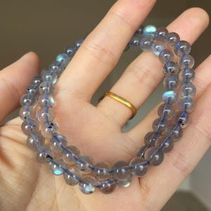 May include: Two strands of round, translucent, gray-blue gemstone beads are displayed. The beads are strung on a blue cord. The beads have a subtle iridescence. A gold ring is visible on a finger in the background. The jewelry is held in a hand.