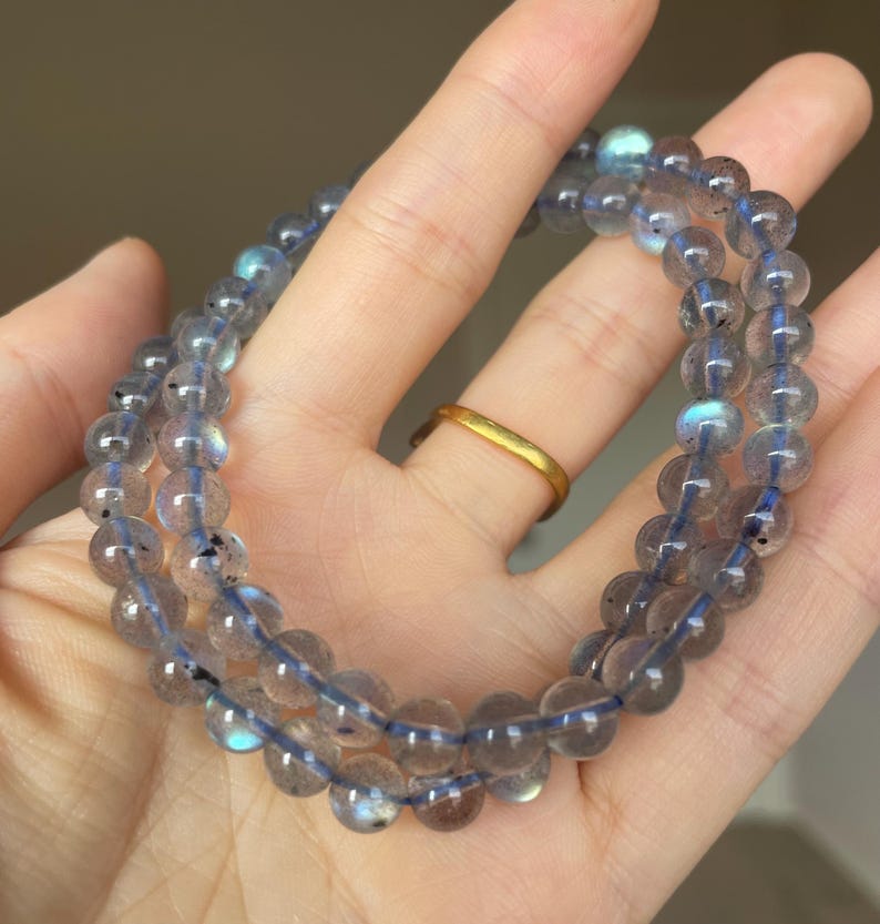 May include: Two strands of round, translucent, gray-blue gemstone beads are displayed. The beads are strung on a blue cord. The beads have a subtle iridescence. A gold ring is visible on a finger in the background. The jewelry is held in a hand.