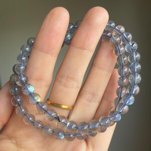 May include: Two beaded bracelets made of translucent, round, gray-blue stones. The beads are strung on a blue cord. The bracelets are held in a hand, with a gold ring on one finger. The background is a neutral, blurred color.