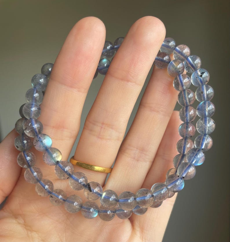 May include: Two beaded bracelets made of translucent, round, gray-blue stones. The beads are strung on a blue cord. The bracelets are held in a hand, with a gold ring on one finger. The background is a neutral, blurred color.