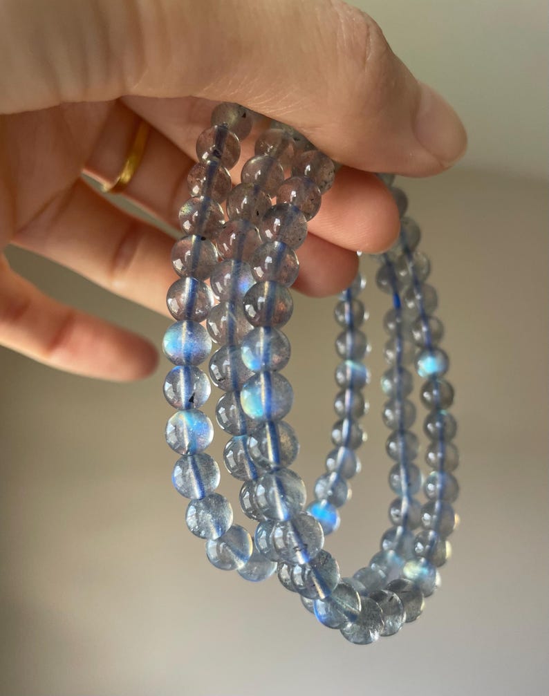 May include: Three light blue beaded bracelets. Each bracelet is made of round, translucent beads with iridescent blue and purple flashes. The bracelets are strung on a thin, clear cord and are held up against a neutral background.