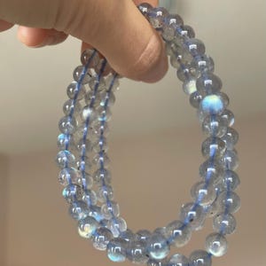 May include: Three strands of beaded bracelets. The beads are a translucent gray with iridescent blue and green flashes. The beads are strung on a blue elastic cord. The bracelets are held up against a light background.