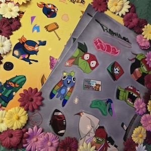May include: Two sticker sheets featuring cartoon characters and designs, surrounded by a floral border. One sheet is yellow with various character illustrations, and the other is gray with more character stickers. The stickers include a variety of colors and shapes.