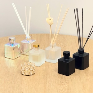 May include: An assortment of decorative reed diffusers and a candle holder on a wooden surface. The diffusers are in various shapes and colors, including iridescent, frosted, and black. Each diffuser has reeds inserted, and some have decorative elements.