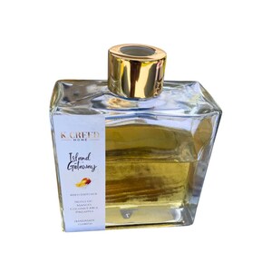 May include: A clear glass bottle with a gold lid and label. The label reads "K Creed Home Island Getaway" and lists notes of mango, coconut milk, and pineapple. The bottle is filled with a yellow liquid.