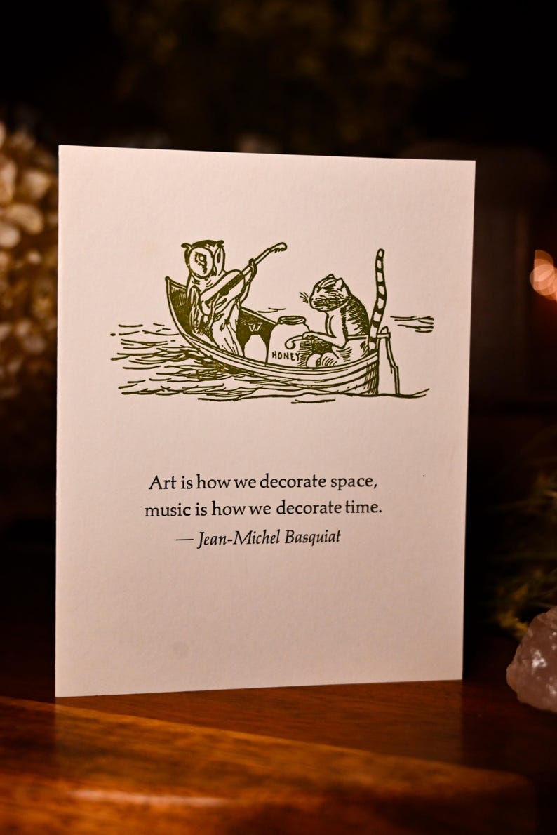 May include: A green and white illustrated card with an owl and a cat in a boat. The text on the card reads "Art is how we decorate space, music is how we decorate time. -Jean-Michel Basquiat"