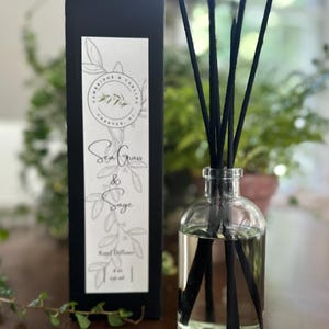May include: A black box with white text that reads "Cambridge & Carling Chester, NJ" and "Sea Grass & Sage Reed Diffuser 6 oz oil". A clear glass bottle with black reeds is filled with a clear liquid. The bottle is sitting on a wooden surface.
