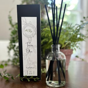 May include: A black box with a white label that says "Cambridge & Carling Chester, NJ" and "Tuscan Olive Reed Diffuser 6 oz 216 ml". The box is next to a clear glass bottle with black reeds and a clear liquid inside.