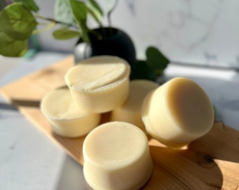 May include: Five round, white, solid, unscented soap bars on a wooden surface. The soap bars are arranged in a pyramid shape.