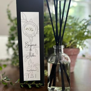 May include: A black box with a white label that reads "Cambridge & Carling, Chester, NJ" and "Bergamot & Amber Reed Diffuser 8 oz / 236 ml". The box is next to a clear glass bottle with black reeds and a clear liquid inside.