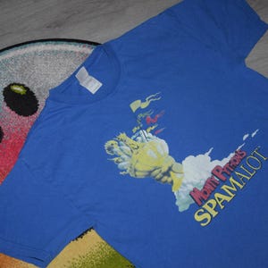 May include: A blue t-shirt with a graphic from Monty Python's Spamalot. The design shows a yellow arm holding a cloud, with the text "Monty Python's SPAMALOT" in yellow and red. The shirt is on a colourful rug.