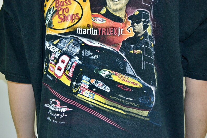 Op de afbeelding: Zwart T-shirt met een NASCAR-raceauto met nummer 8 en de woorden "Bass Pro Shops" en "Martin Truex Jr." Het shirt heeft ook de woorden "Monte Carlo" en diverse sponsorlogo's.