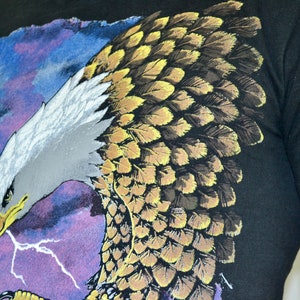 Może przedstawiać: Czarny t-shirt z szczeg&oacute;łową grafiką orła. Orzeł ma białą głowę, ż&oacute;łty dzi&oacute;b oraz brązowe i ż&oacute;łte pi&oacute;ra. Tło przedstawia niebiesko-fioletowe niebo z białymi błyskawicami.