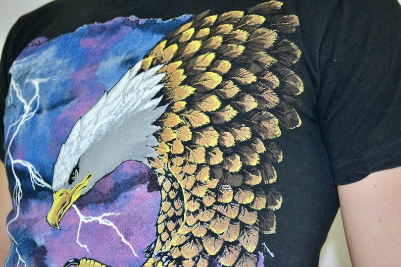 Może przedstawiać: Czarny t-shirt z szczeg&oacute;łową grafiką orła. Orzeł ma białą głowę, ż&oacute;łty dzi&oacute;b oraz brązowe i ż&oacute;łte pi&oacute;ra. Tło przedstawia niebiesko-fioletowe niebo z białymi błyskawicami.