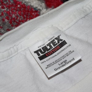 May include: White cotton t-shirt tag with black text that reads "TULTEX", "COTTON", "100% COTTON/ALGOD&Oacute;N/COTON", "MADE IN MEXICO/HECHO EN MEXICO/FABRIQUE AUX MEXIQUE", "Large", and "Grande/Grand".