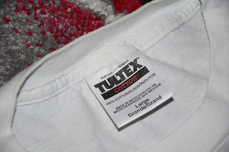 May include: White cotton t-shirt tag with black text that reads "TULTEX", "COTTON", "100% COTTON/ALGOD&Oacute;N/COTON", "MADE IN MEXICO/HECHO EN MEXICO/FABRIQUE AUX MEXIQUE", "Large", and "Grande/Grand".