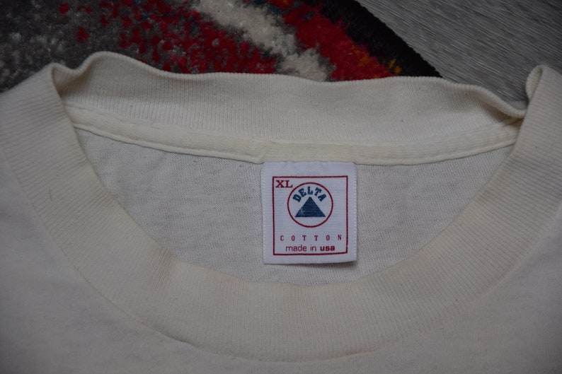 May include: White cotton t-shirt tag with a white triangle logo that says "Delta" and "Cotton made in USA". The tag also says "XL".