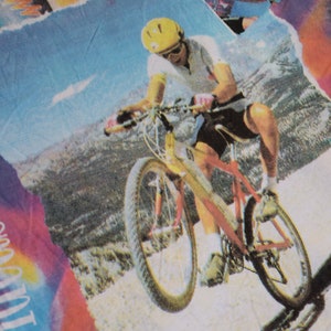 Puede incluir: Una persona con un casco amarillo y una camisa blanca monta una bicicleta de monta&ntilde;a roja y plateada en un camino de tierra. El fondo es un cielo azul con nubes blancas y una cordillera.