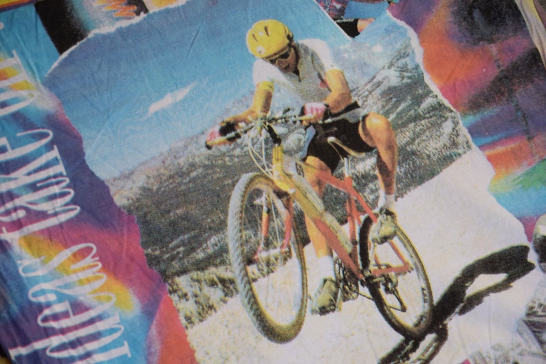 Puede incluir: Una persona con un casco amarillo y una camisa blanca monta una bicicleta de monta&ntilde;a roja y plateada en un camino de tierra. El fondo es un cielo azul con nubes blancas y una cordillera.