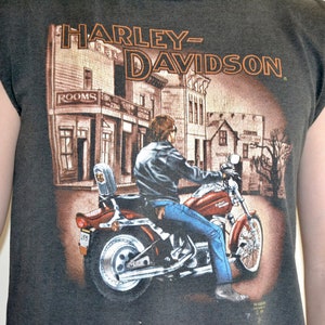 May include: Dark gray sleeveless t-shirt featuring a Harley-Davidson graphic. The graphic depicts a person riding a red motorcycle in front of a town scene. The Harley-Davidson logo is displayed prominently at the top.