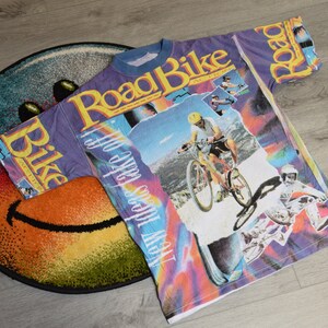 Puede incluir: Una camiseta morada, azul y blanca con un estampado gr&aacute;fico de un ciclista de monta&ntilde;a y el texto "Road Bike" y "How 'bout we take off".