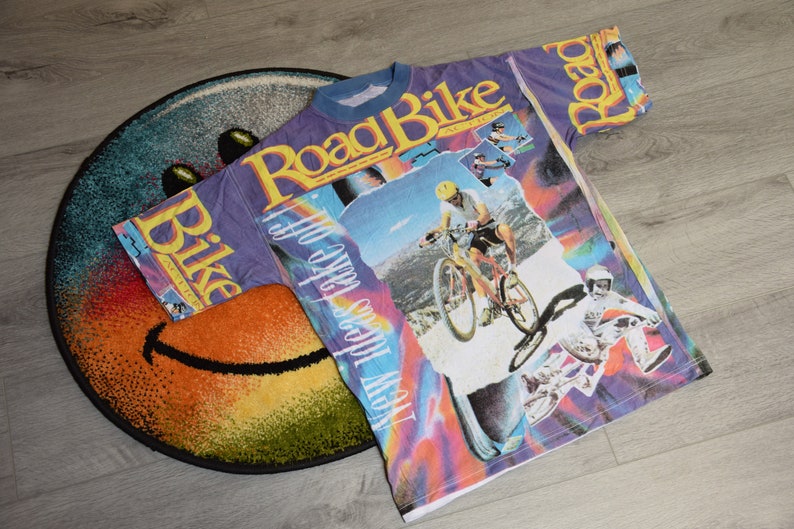 Puede incluir: Una camiseta morada, azul y blanca con un estampado gr&aacute;fico de un ciclista de monta&ntilde;a y el texto "Road Bike" y "How 'bout we take off".