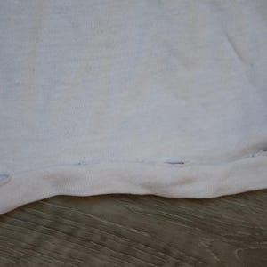 Op de afbeelding: Close-up van een wit stoffen kledingstuk, mogelijk een t-shirt of iets dergelijks. De zoom aan de onderkant is zichtbaar, met een naad met enkele kleine gaatjes. De stof lijkt enigszins versleten.