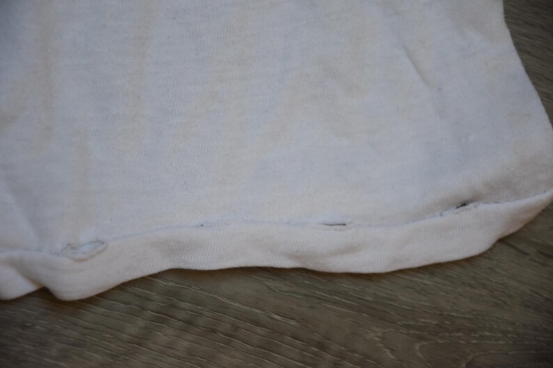 Op de afbeelding: Close-up van een wit stoffen kledingstuk, mogelijk een t-shirt of iets dergelijks. De zoom aan de onderkant is zichtbaar, met een naad met enkele kleine gaatjes. De stof lijkt enigszins versleten.